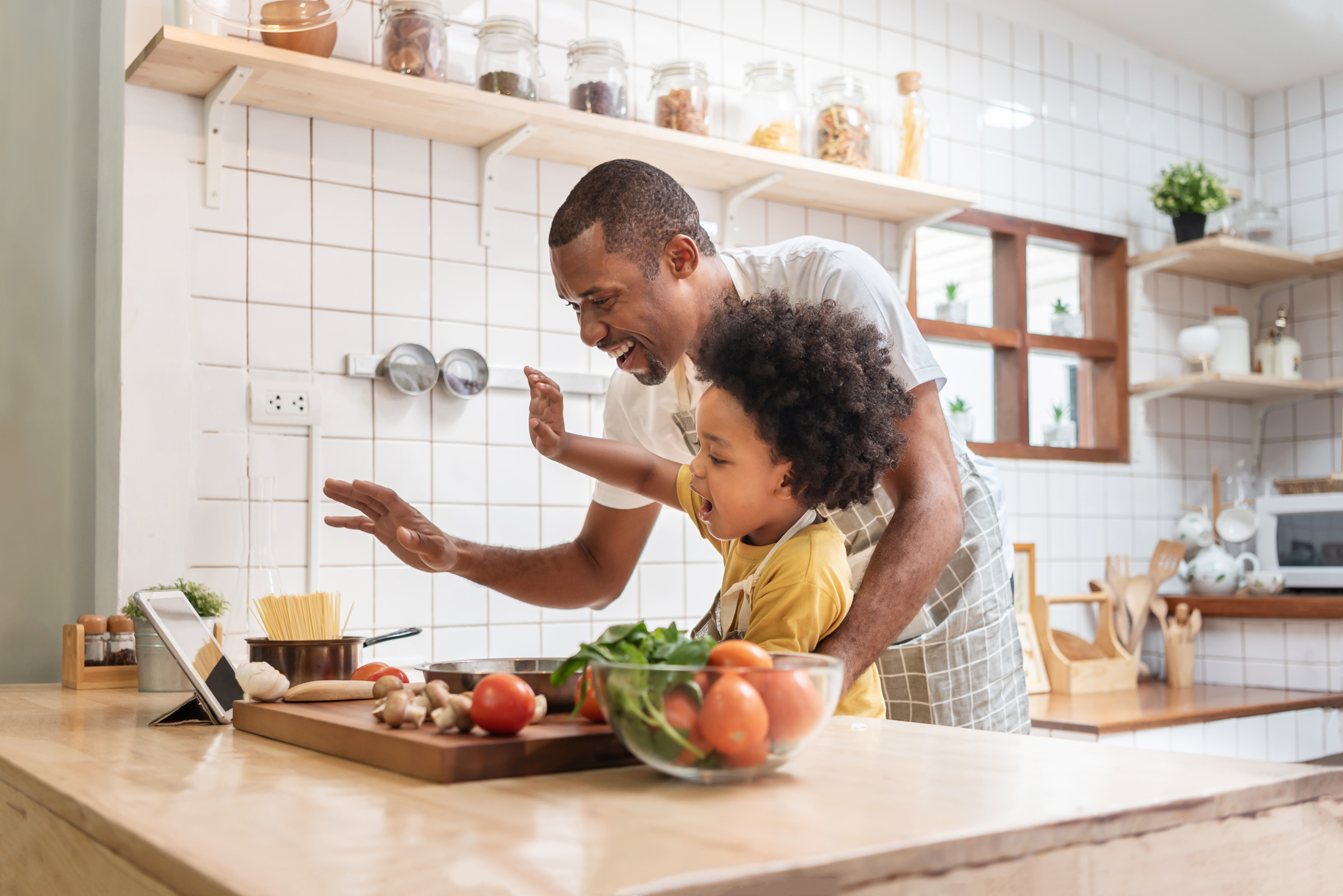 Man and young child waving at a tablet while cooking together in a kitchen.