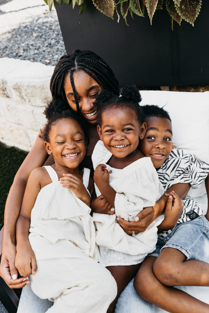woman hugging three kids outdoors Smiling woman with braids hugging three laughing young children on an outdoor bench, two girls in white outfits and a boy in a patterned shirt.